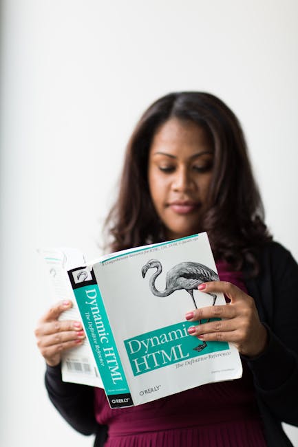 A woman with long dark hair, wearing a black blazer over a burgundy top, stands indoors against a plain white wall, holding a book titled 'Dynamic HTML' with a cover illustration of a flamingo. She ap