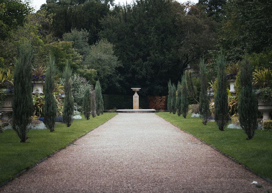 A lush garden scene featuring a neatly mowed lawn with vibrant green grass, bordered by well-maintained garden beds filled with flowering plants and shrubs. In the background, a row of tall, trimmed h